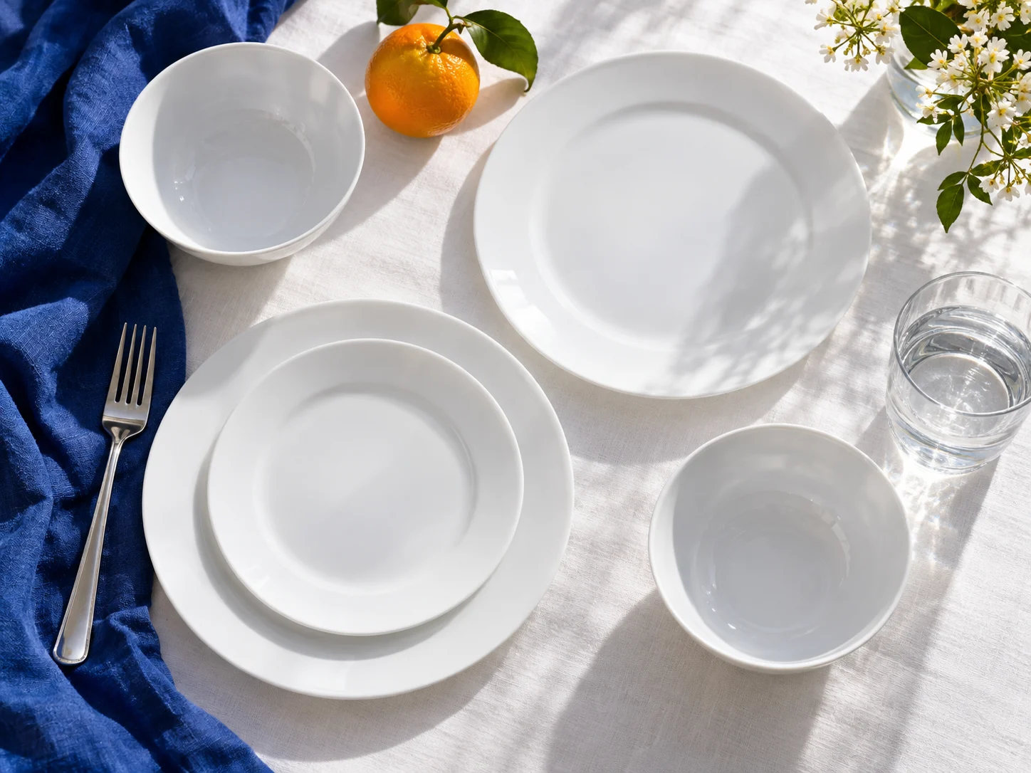 Illustrative thin white plates and bowls on a white table with cobalt linen and an orange